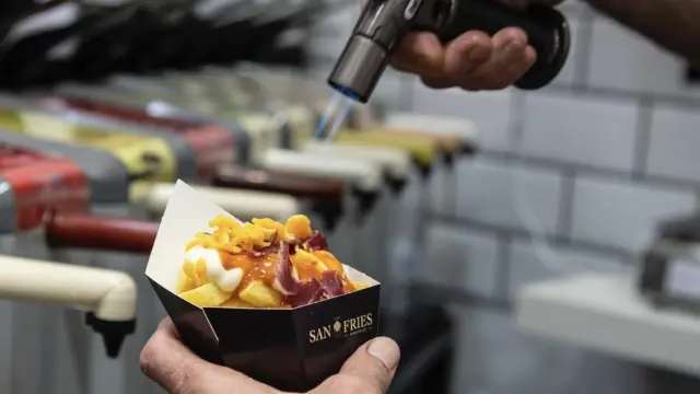 Una persona sosteniendo un cono de patatas fritas con salsas con una mano y, con la otra, un soplete de cocina
