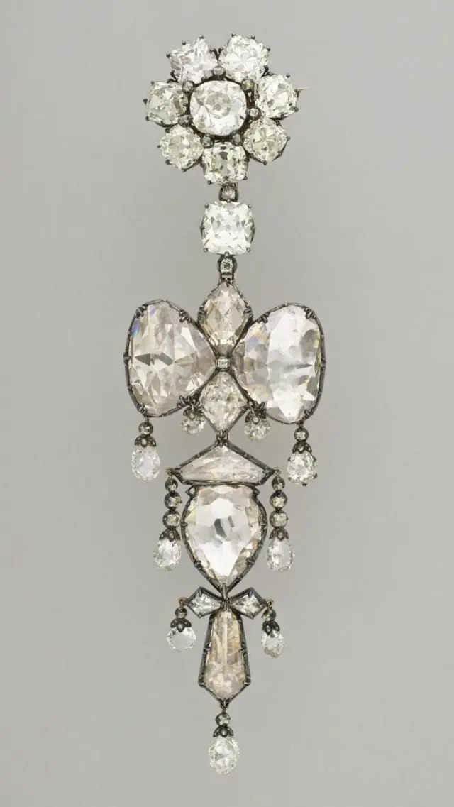Roseta formada por siete diamantes que rodean un solitario. En total el conjunto está compuesto por 92 diamantes de diferentes tamaños. Su orfebre se inspiró en un modelo de plomo del siglo XVIII.