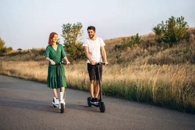 Dos jóvenes circulan en patinete eléctrico por un camino asfaltado.